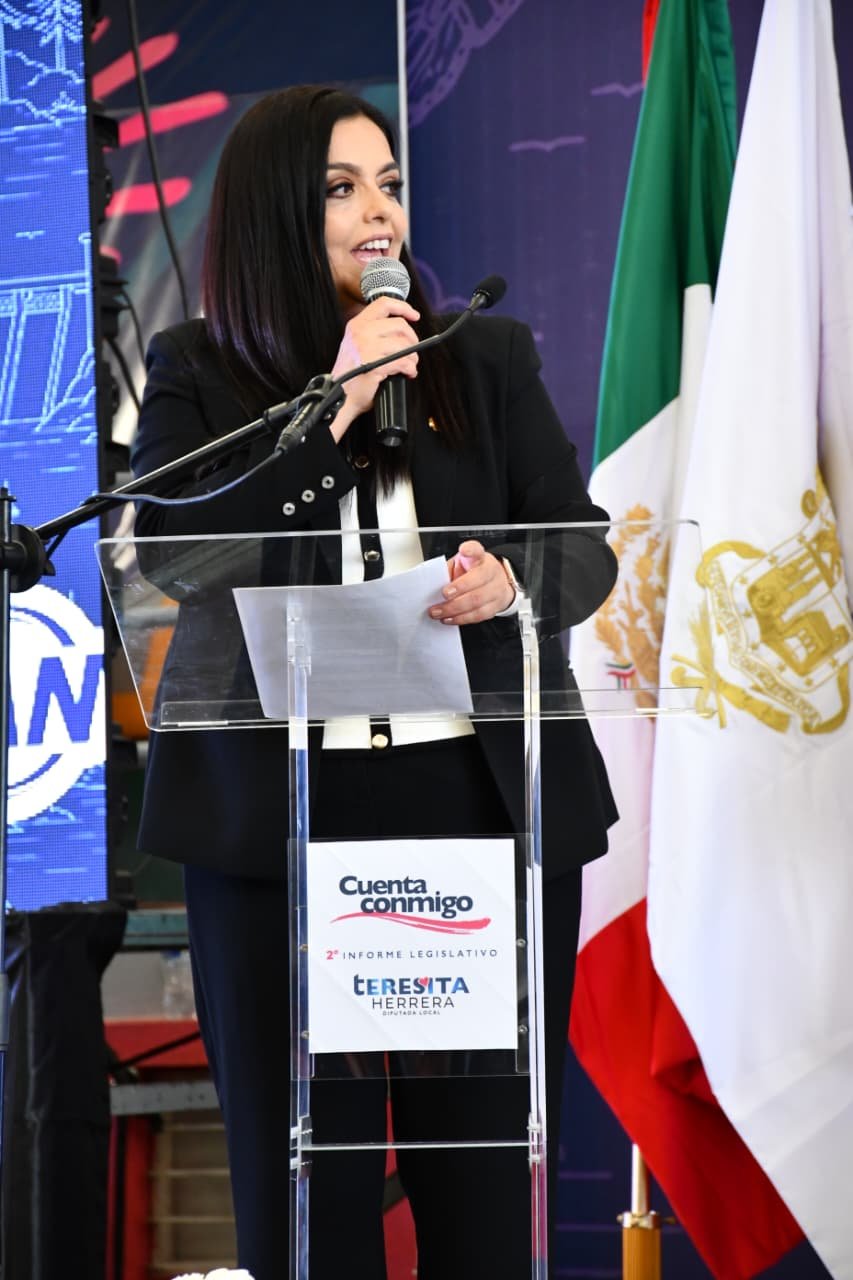 Tere Herrera hablando ante micrófonos y bandera de Michoacán durante su segundo informe legislativo en el Congreso del